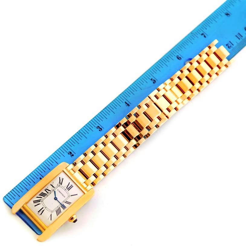 The image shows a top-down view of the Cartier Tank Americaine 1735 Men
s Yellow Gold Silver Dial 1735 Men
s Yellow Gold Silver Dial watch, highlighting its face, bezel, and metal bracelet against a measuring ruler.