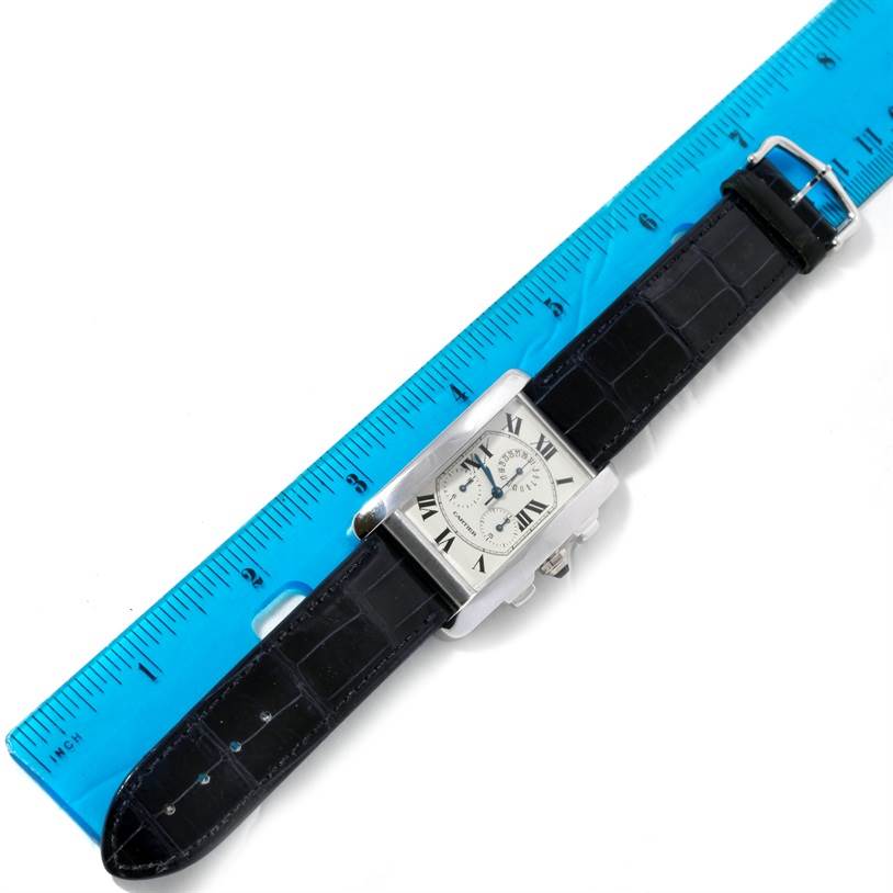 The Cartier Tank Americaine W2603358 White Gold Silver Dial W2603358 White Gold Silver Dial watch is shown from a top view, laying against a ruler, displaying its face and black leather strap.