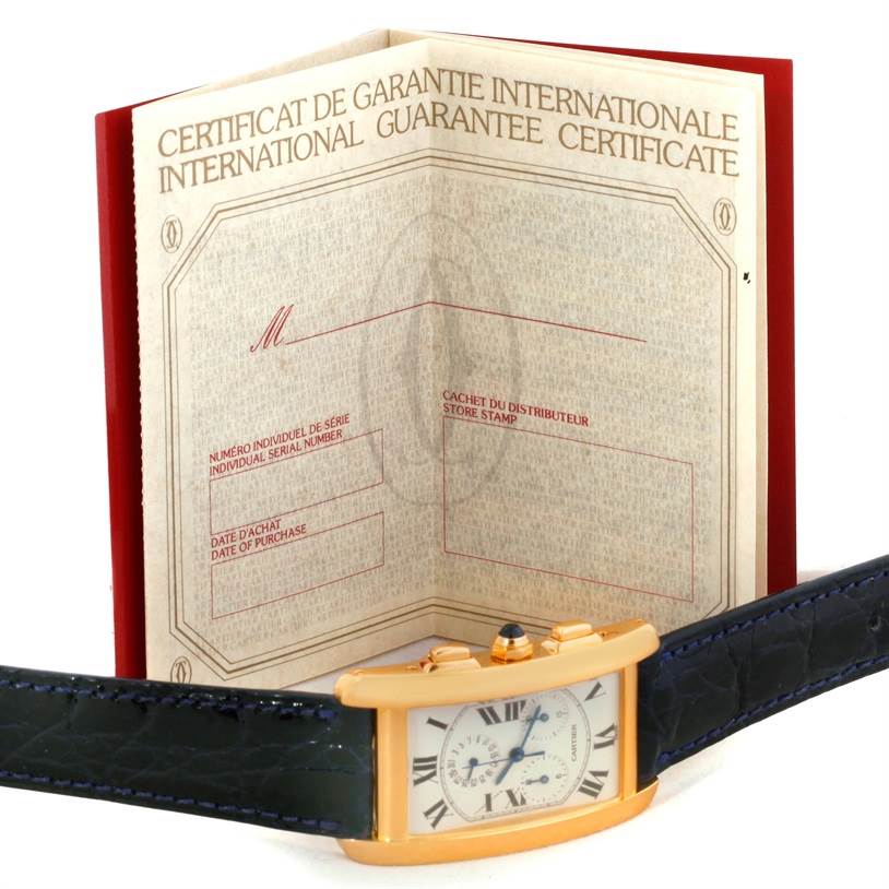 The image shows a Cartier Tank Americaine W2601156 Men
s Yellow Gold Silver Dial W2601156 Men
s Yellow Gold Silver Dial watch with a black leather strap, placed in front of an open international guarantee certificate.