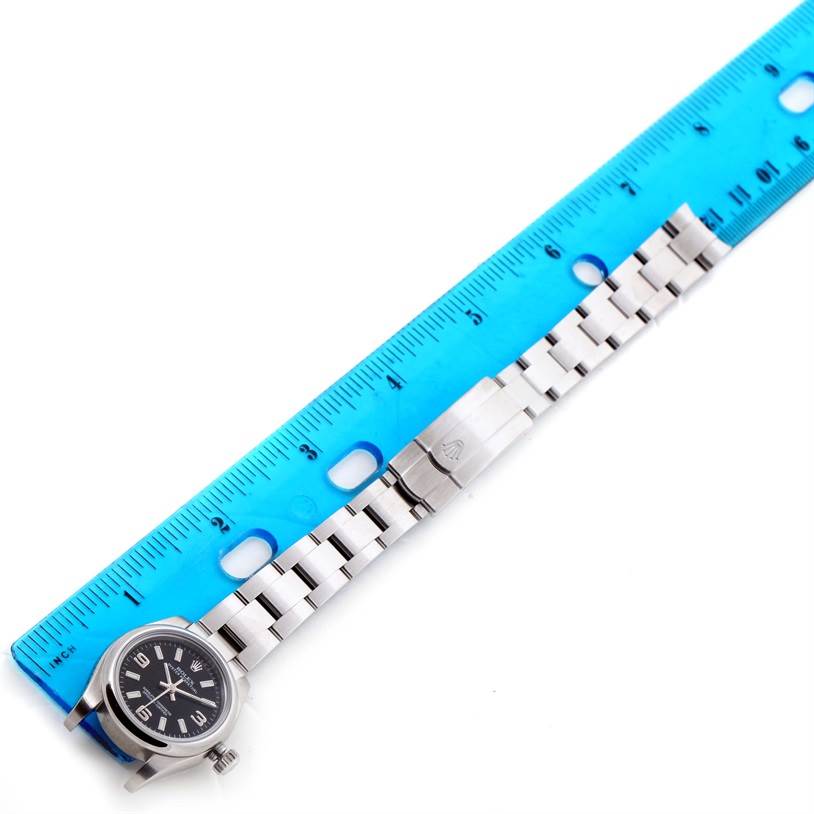 The image shows an Oyster Perpetual Rolex watch laid alongside a blue ruler, displaying the watch face and bracelet.
