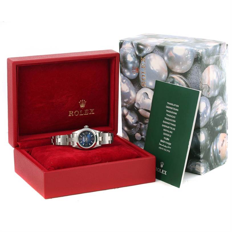 The Rolex Oyster Perpetual 76080 Women
s Stainless Steel Blue Dial 76080 Women
s Stainless Steel Blue Dial watch is displayed frontally in its red box, along with its packaging and instruction manual.