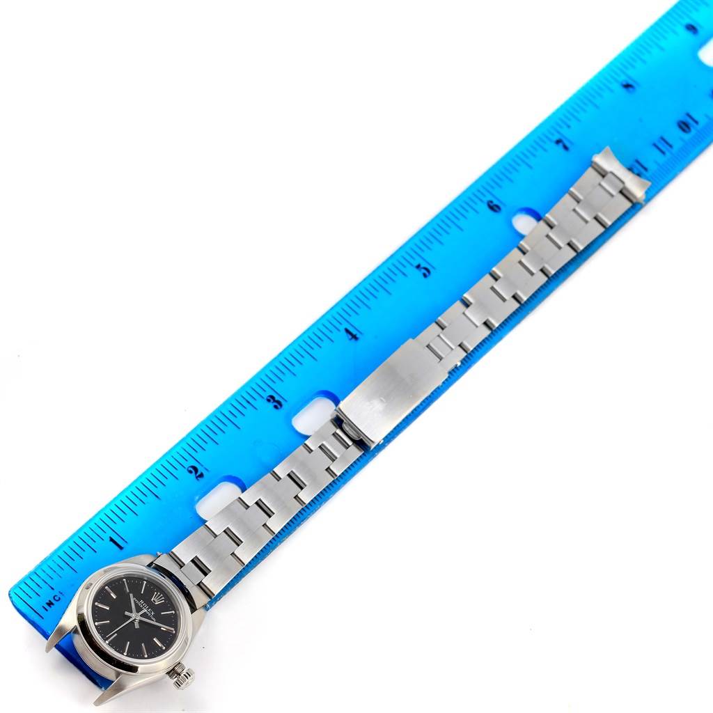 The Rolex Oyster Perpetual 76080 Women's Stainless Steel Black Dial 76080 Women's Stainless Steel Black Dial watch is shown from a top-down angle alongside a blue ruler, highlighting its bracelet and clasp.