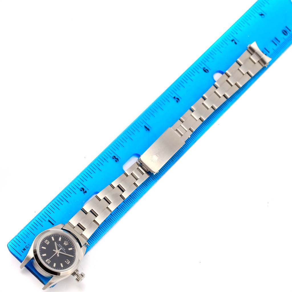 The image shows a Rolex Oyster Perpetual 67180 Women's Stainless Steel Black Dial 67180 Women's Stainless Steel Black Dial watch laid flat next to a blue ruler, showcasing its face, bracelet, and clasp.