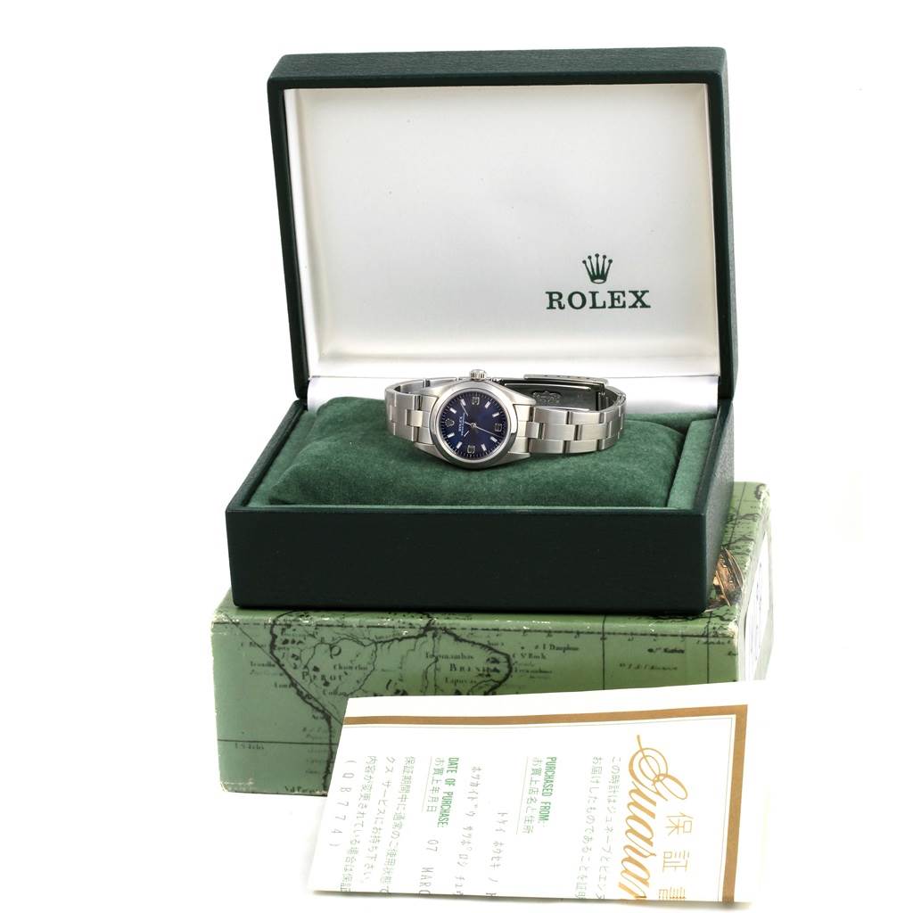 The Rolex Oyster Perpetual 76080 Women
s Stainless Steel Blue Dial 76080 Women
s Stainless Steel Blue Dial watch is shown front-facing in its open box, atop green cushioning, with the brand logo visible.