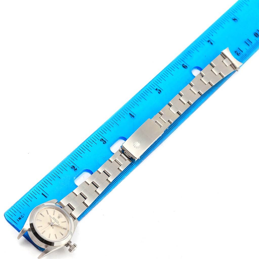 The image shows a Rolex Oyster Perpetual 67180 Women
s Stainless Steel Silver Dial 67180 Women
s Stainless Steel Silver Dial watch and band, with the band laid out alongside a ruler.