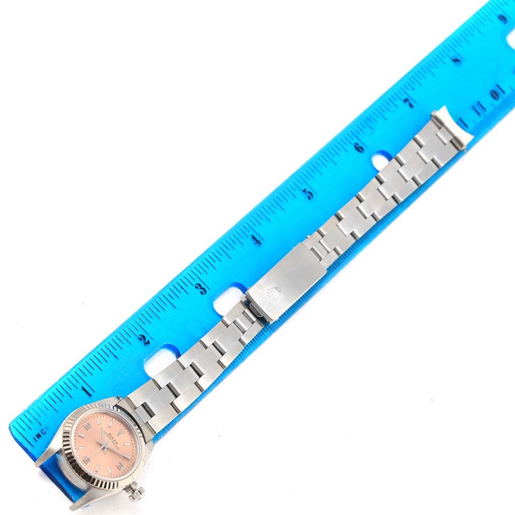 The image shows a Rolex Oyster Perpetual 76094 Women
s Stainless Steel Pink Dial 76094 Women
s Stainless Steel Pink Dial watch placed next to a blue ruler, highlighting the watch face and metal bracelet.