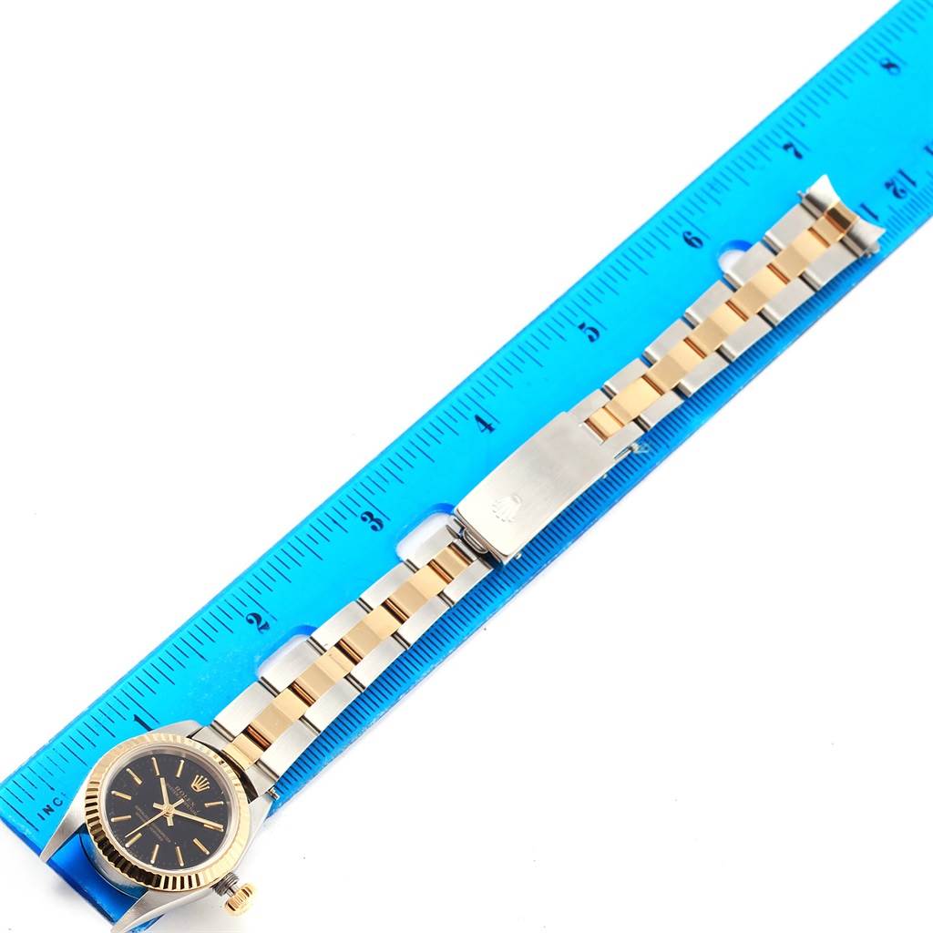 The Rolex Oyster Perpetual 67193 Women's Steel and Gold (two tone) Black Dial 67193 Women's Steel and Gold (two tone) Black Dial watch is shown from a top-down angle, along with a blue ruler for size reference.