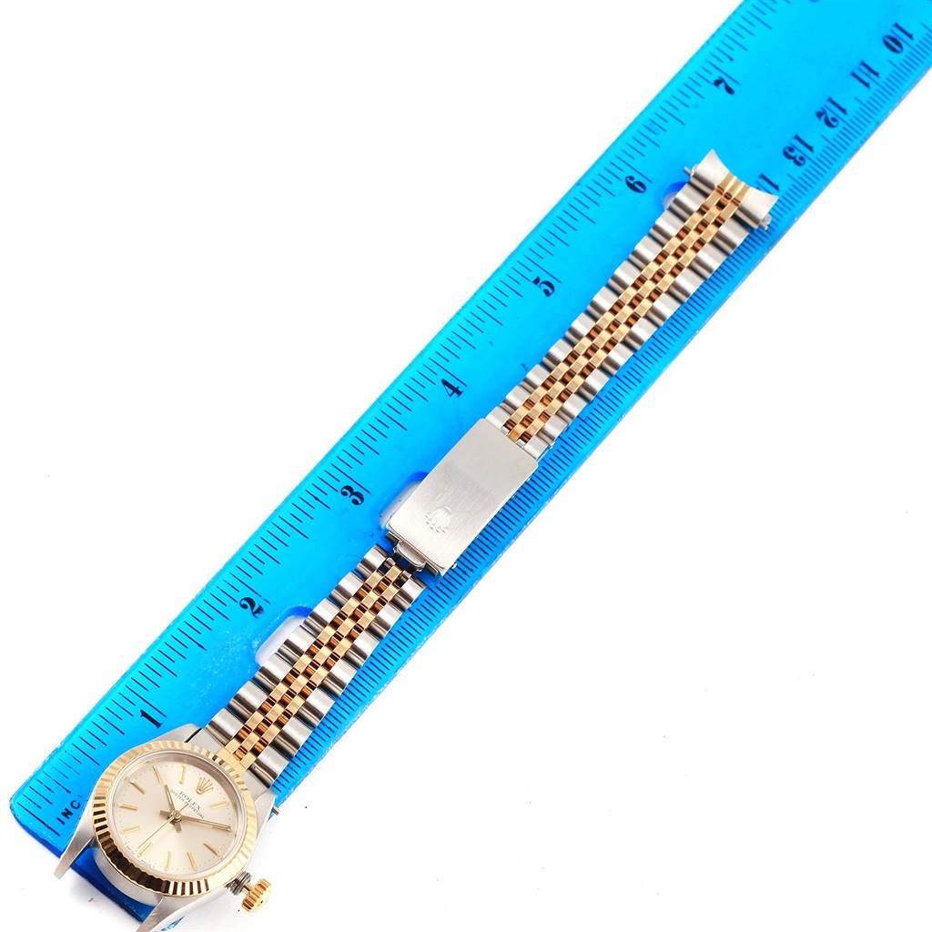 The image shows an overhead view of a Rolex Oyster Perpetual 67193 Women
s Steel and Gold (two tone) Silver Dial 67193 Women
s Steel and Gold (two tone) Silver Dial watch with a two-tone metal bracelet aligned next to a blue ruler.