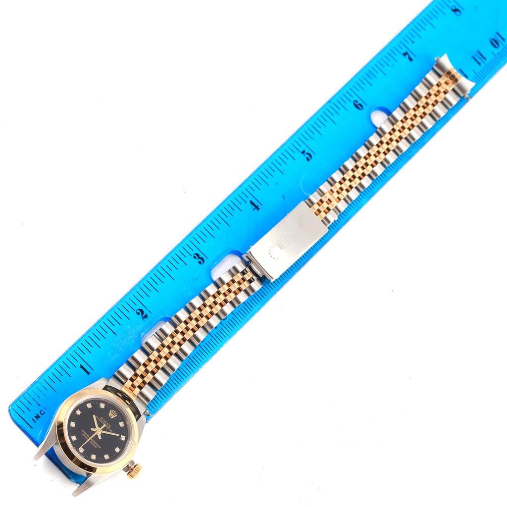 The image shows a Rolex Oyster Perpetual 76183 Women's Steel and Gold (two tone) Black Dial 76183 Women's Steel and Gold (two tone) Black Dial watch face and bracelet arranged beside a blue ruler.