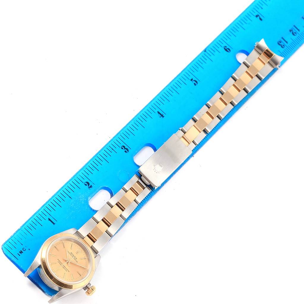 The image shows a Rolex Oyster Perpetual 76183 Women
s Steel and Gold (two tone) Champagne Dial 76183 Women
s Steel and Gold (two tone) Champagne Dial watch laid flat next to a blue ruler, highlighting its bracelet and dial.