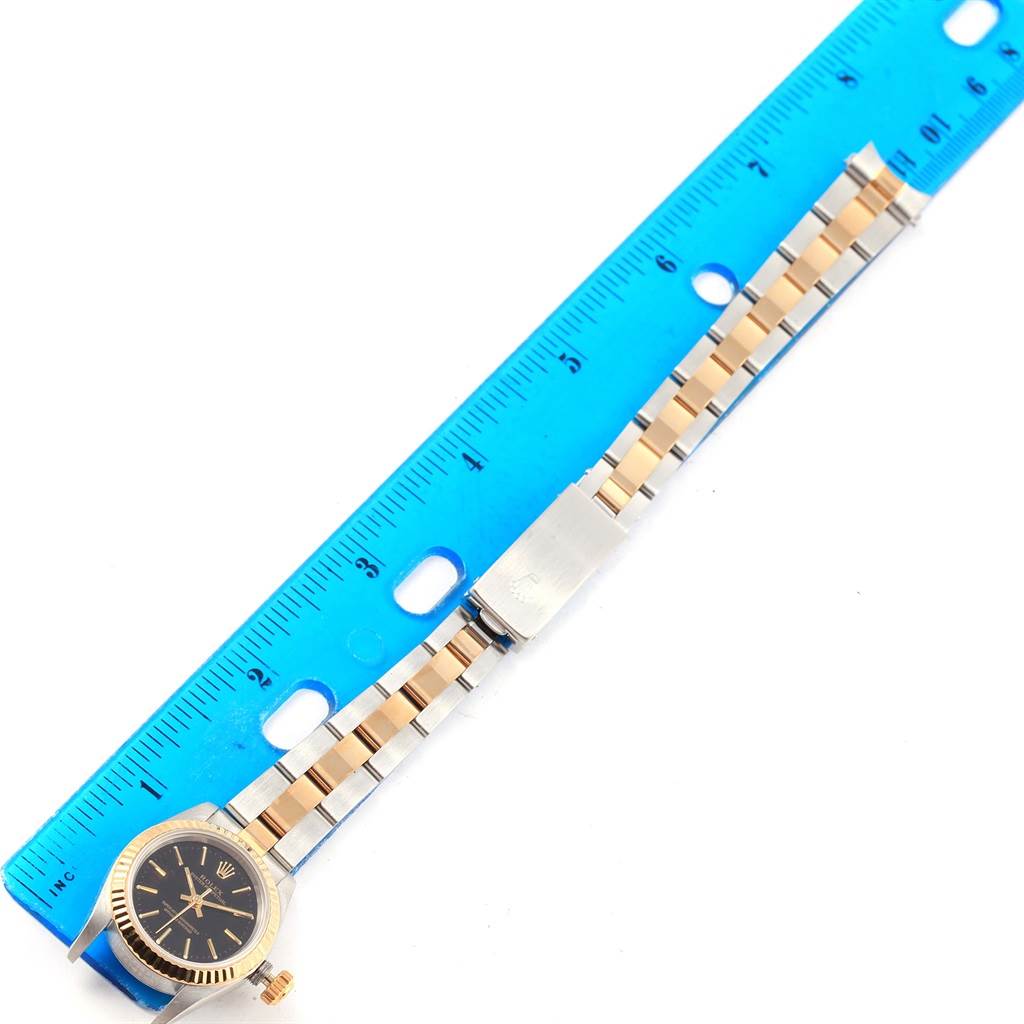 The image shows a Rolex Oyster Perpetual 67193 Women's Steel and Gold (two tone) Black Dial 67193 Women's Steel and Gold (two tone) Black Dial watch with the face and band aligned next to a blue ruler for measurement.