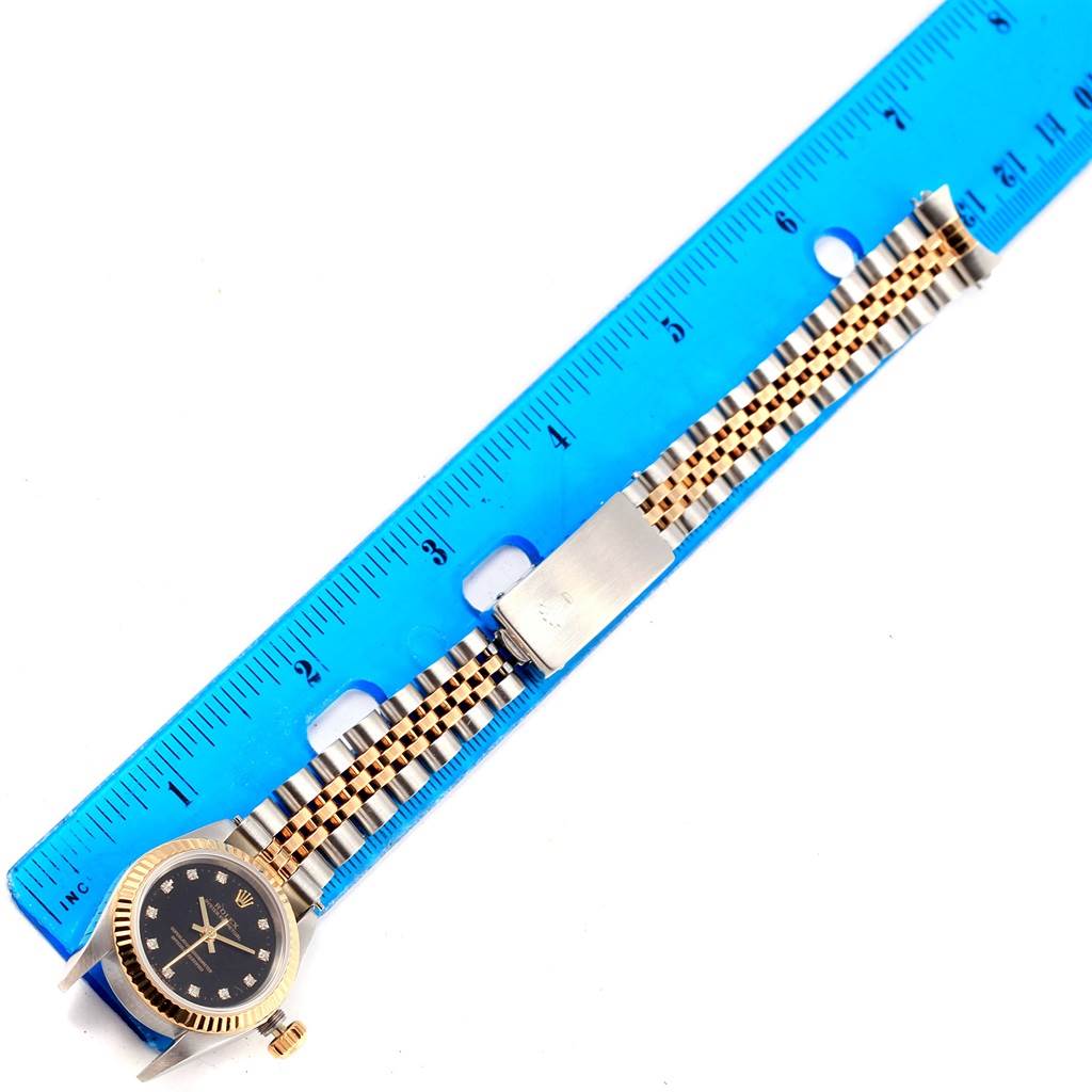 The image shows the Rolex Oyster Perpetual 67193 Women's Steel and Gold (two tone) Black Dial 67193 Women's Steel and Gold (two tone) Black Dial watch placed next to a blue ruler, highlighting the watch face, band, and clasp.