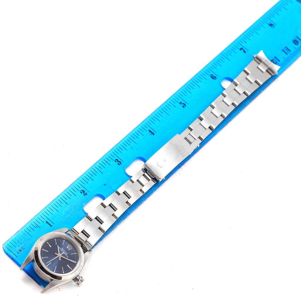 The image shows a Rolex Oyster Perpetual 76080 Women's Stainless Steel Blue Dial 76080 Women's Stainless Steel Blue Dial watch laid flat on a blue ruler, displaying its face, bracelet, and clasp.