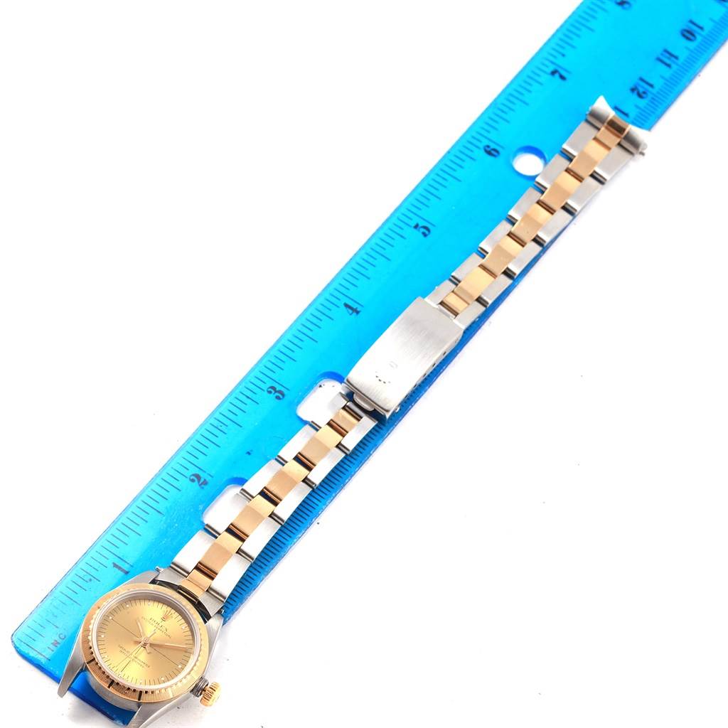 The image shows a Rolex Oyster Perpetual 76243 Women's Steel and Gold (two tone) Champagne Dial 76243 Women's Steel and Gold (two tone) Champagne Dial watch laid out flat beside a ruler, displaying its gold-and-silver bracelet and face.