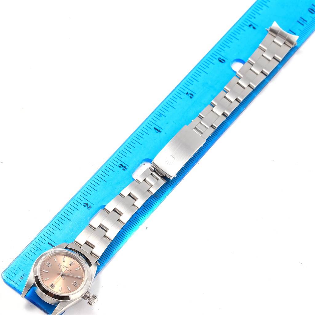 The image shows a Rolex Oyster Perpetual 67180 Women
s Stainless Steel Pink Dial 67180 Women
s Stainless Steel Pink Dial watch with a metal bracelet and clasp, aligned next to a blue ruler.