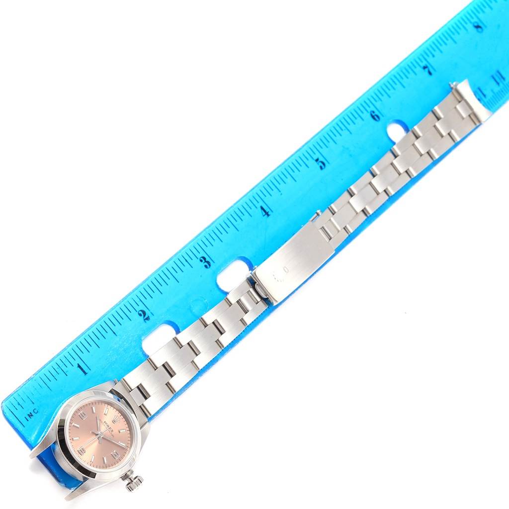 The image displays an overhead view of a Rolex Oyster Perpetual 67180 Women
s Stainless Steel Pink Dial 67180 Women
s Stainless Steel Pink Dial watch on a blue ruler, showing the face and stainless steel bracelet fully extended.