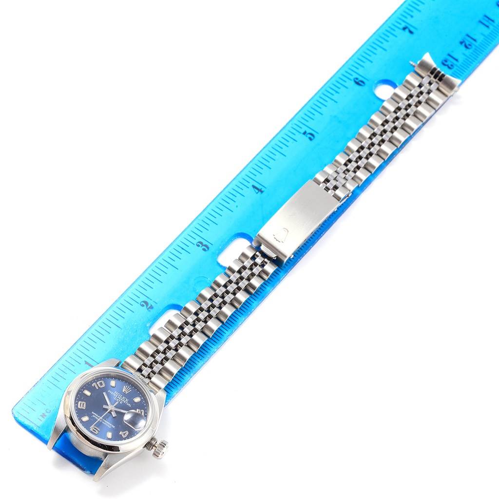 The image shows a Rolex Oyster Perpetual 79160 Women's Stainless Steel Blue Dial 79160 Women's Stainless Steel Blue Dial watch laid flat next to a blue ruler, displaying its face, bracelet, and clasp.