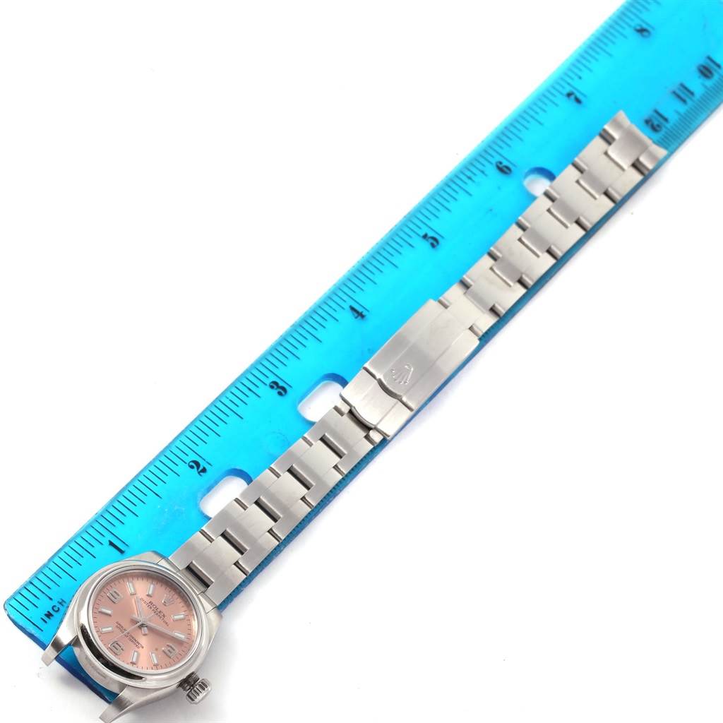 The image shows a Rolex Oyster Perpetual 176200 Women
s Stainless Steel Pink Dial 176200 Women
s Stainless Steel Pink Dial watch with a pink dial and stainless steel bracelet alongside a blue ruler.