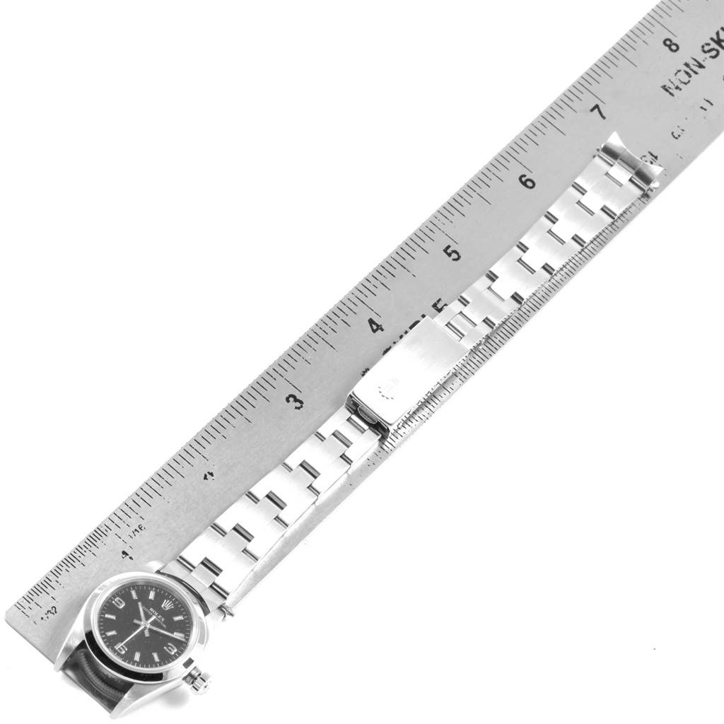 The Rolex Oyster Perpetual 76080 Women's Stainless Steel Black Dial 76080 Women's Stainless Steel Black Dial watch is shown from an overhead angle, displaying the face, band, and clasp next to a ruler.
