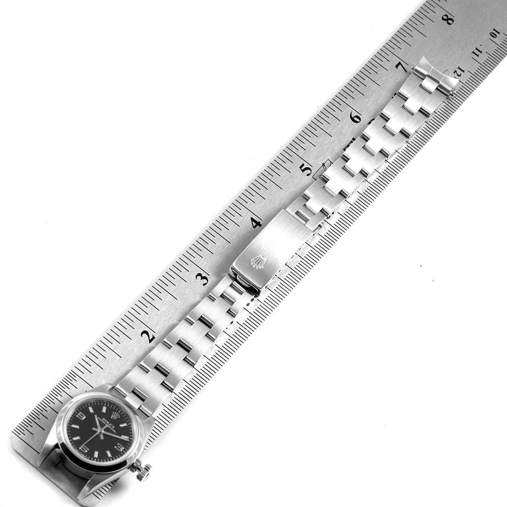 The image shows a top view of a Rolex Oyster Perpetual 67180 Women
s Stainless Steel Black Dial 67180 Women
s Stainless Steel Black Dial watch, featuring its bezel, dial, bracelet, and clasp, alongside a ruler.
