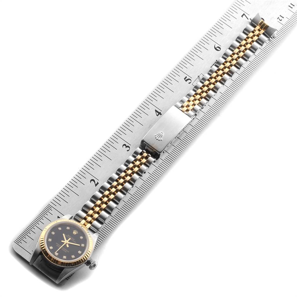 The image shows the full length of a Rolex Oyster Perpetual 76193 Women
s Steel and Gold (two tone) Black Dial 76193 Women
s Steel and Gold (two tone) Black Dial watch laid flat on a metal ruler.