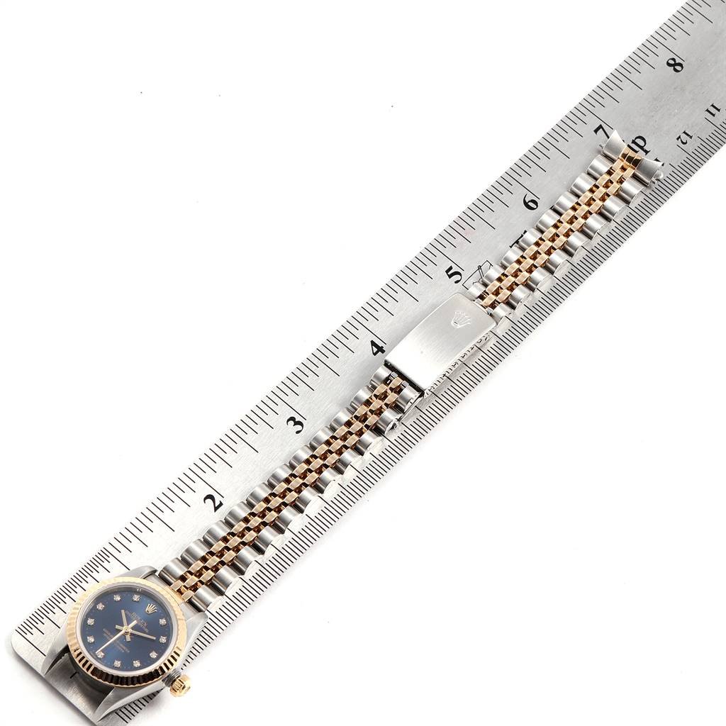 The image shows a Rolex Oyster Perpetual 76193 Women
s Steel and Gold (two tone) Blue Dial 76193 Women
s Steel and Gold (two tone) Blue Dial watch laid flat on a ruler, displaying the face, bracelet, and clasp.
