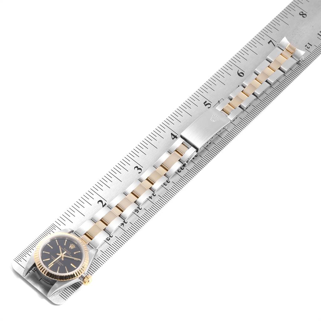 The image shows the Rolex Oyster Perpetual 76193 Women
s Steel and Gold (two tone) Black Dial 76193 Women
s Steel and Gold (two tone) Black Dial watch laid flat with a close-up angle displaying its face and bracelet against a ruler.