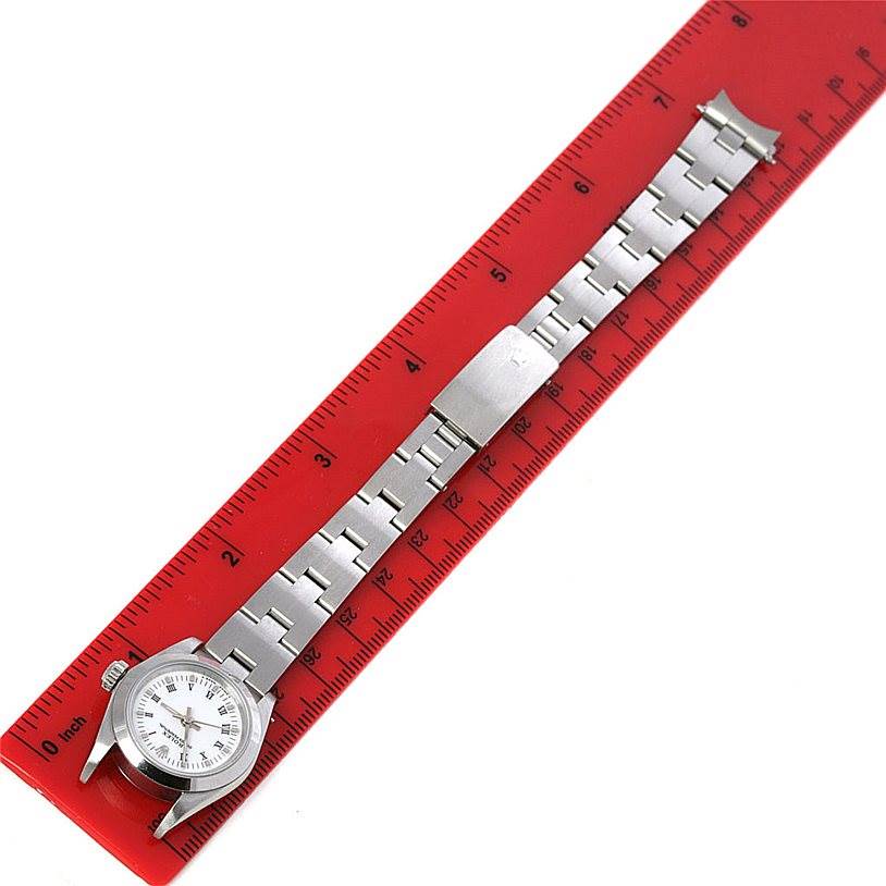 The image shows the Rolex Oyster Perpetual 67180 Women's Stainless Steel White Dial 67180 Women's Stainless Steel White Dial watch laid flat on a red ruler, displaying the watch face and metal bracelet.