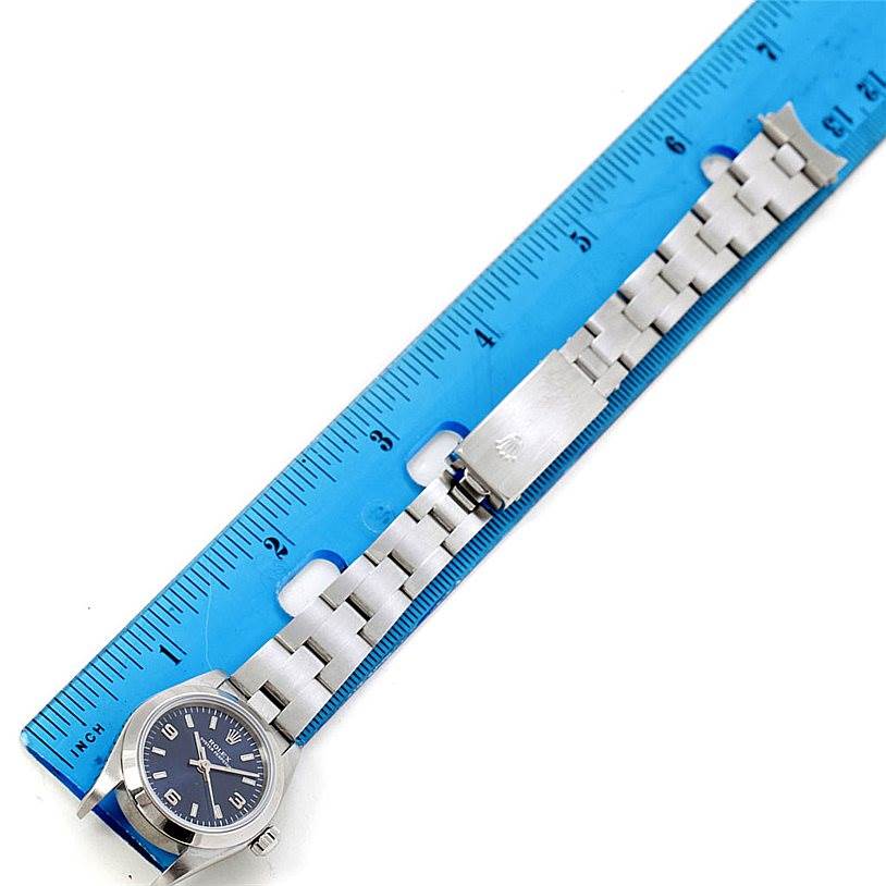 The image shows an aerial view of a Rolex Oyster Perpetual 76080 Women's Stainless Steel Blue Dial 76080 Women's Stainless Steel Blue Dial watch alongside a blue ruler, displaying the watchband and clasp.