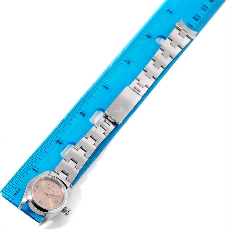 The Rolex Oyster Perpetual 76080 Women's Stainless Steel Pink Dial 76080 Women's Stainless Steel Pink Dial watch is shown horizontally with its bracelet next to a blue ruler, displaying the face, crown, and clasp.