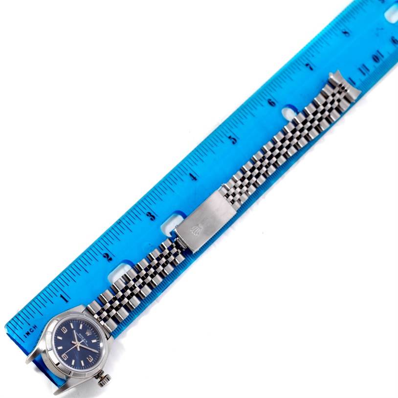 The Rolex Oyster Perpetual 67230 Women's Stainless Steel Blue Dial 67230 Women's Stainless Steel Blue Dial watch is shown from above, highlighting the face, bracelet, and clasp next to a blue ruler for scale.