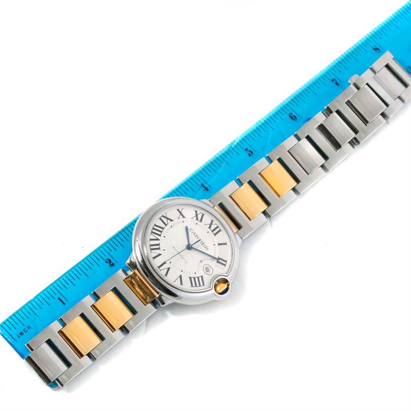 The image shows the Ballon Bleu model by Cartier placed on a ruler, displaying its face and metal bracelet.