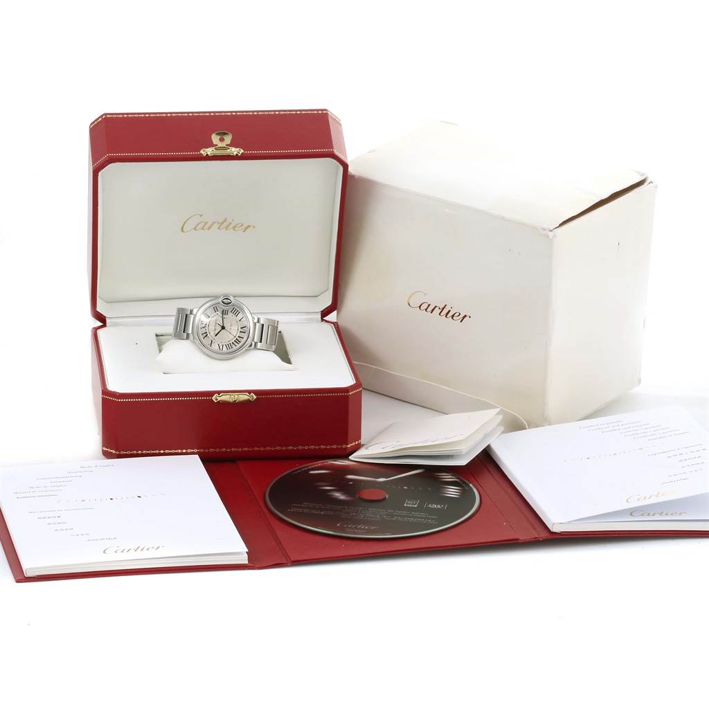 The image shows a Cartier Ballon Bleu W6920046 Men
s Stainless Steel Silver Dial W6920046 Men
s Stainless Steel Silver Dial watch in an open red box, with accompanying packaging, booklet, and a CD.