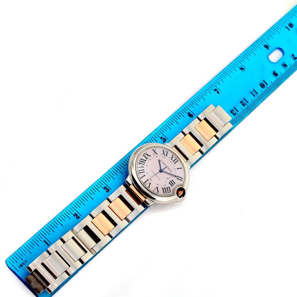 The image shows a Cartier Ballon Bleu W6920070 Women's Steel and Gold (two tone) Mother of Pearl Dial W6920070 Women's Steel and Gold (two tone) Mother of Pearl Dial watch flat, next to a blue ruler, highlighting its band and watch face.