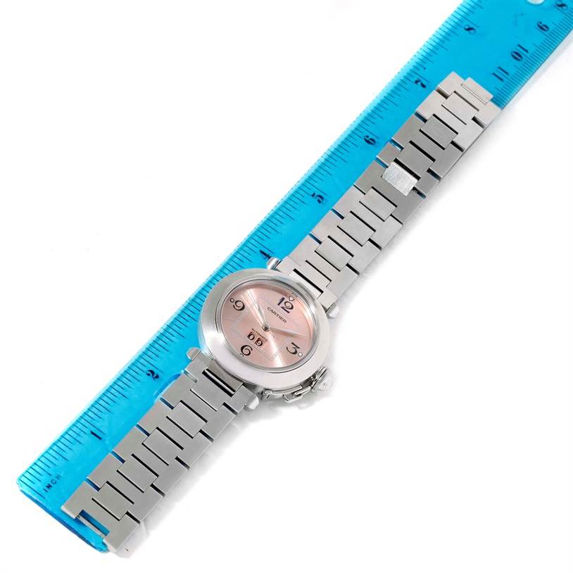 The Cartier Pasha W31058M7 Women
s Stainless Steel Pink Dial W31058M7 Women
s Stainless Steel Pink Dial watch is shown from a top-down angle, featuring its face, bezel, and metal bracelet alongside a ruler.