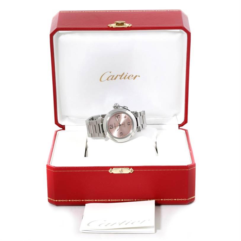 The Cartier Pasha W31075M7 Women
s Stainless Steel Pink Dial W31075M7 Women
s Stainless Steel Pink Dial watch is shown front-facing in an open box along with paperwork beneath the box.