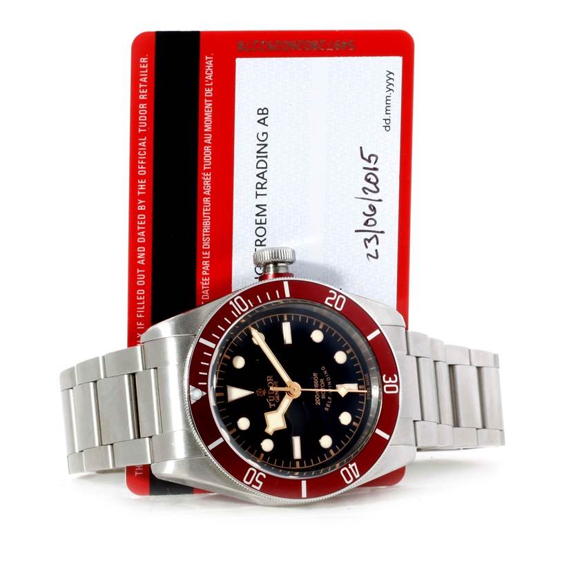 The Tudor Heritage Black Bay 79220R Men's Stainless Steel Black Dial 79220R Men's Stainless Steel Black Dial watch is shown from the front, with the stainless steel bracelet and dial visible, along with its warranty card in the background.