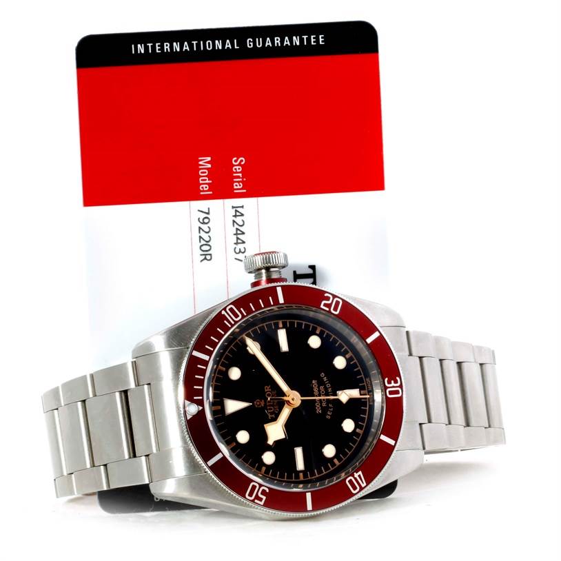 The Tudor Heritage Black Bay 79220R Men's Stainless Steel Black Dial 79220R Men's Stainless Steel Black Dial watch is shown at a slightly tilted angle, displaying the dial, bezel, bracelet, and a warranty card in the background.