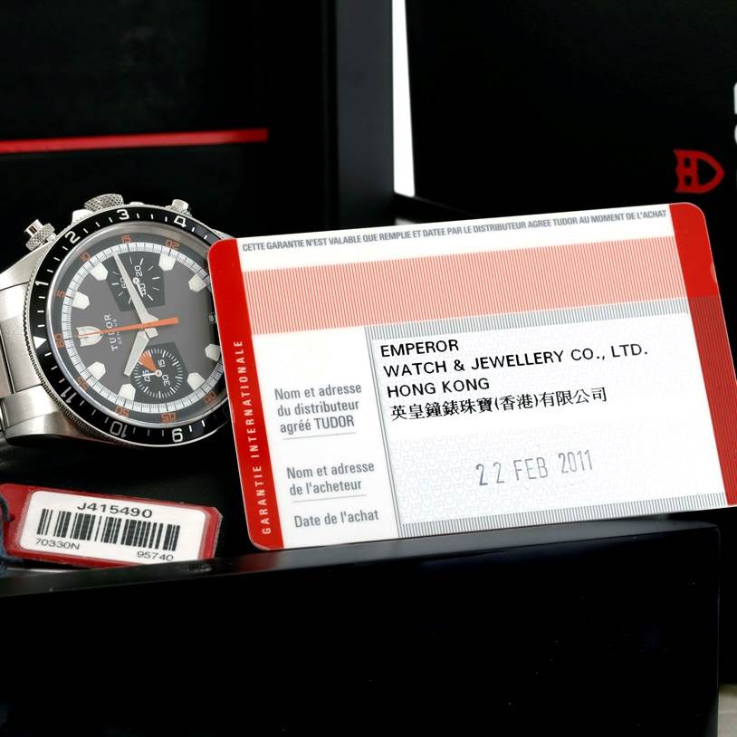 The image shows a Tudor Heritage Chrono 70330N-95740 Men's Stainless Steel Grey Dial 70330N-95740 Men's Stainless Steel Grey Dial watch viewed from a slight side angle, along with its warranty card and barcode.
