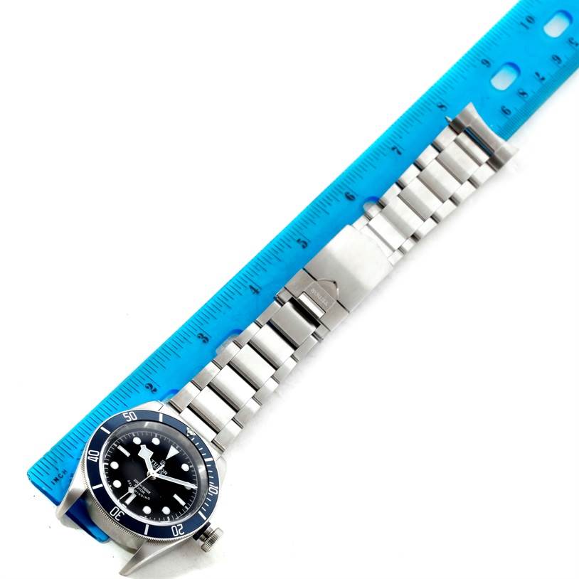 The image shows a top view of a Tudor Heritage Black Bay 79220 Men's Stainless Steel Black Dial 79220 Men's Stainless Steel Black Dial watch with its entire stainless steel bracelet laid beside a blue ruler.
