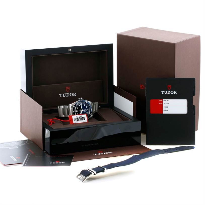 The Tudor Heritage Black Bay 79220 Men's Stainless Steel Black Dial 79220 Men's Stainless Steel Black Dial watch is displayed at an angled view in its open box with documents and an additional strap.