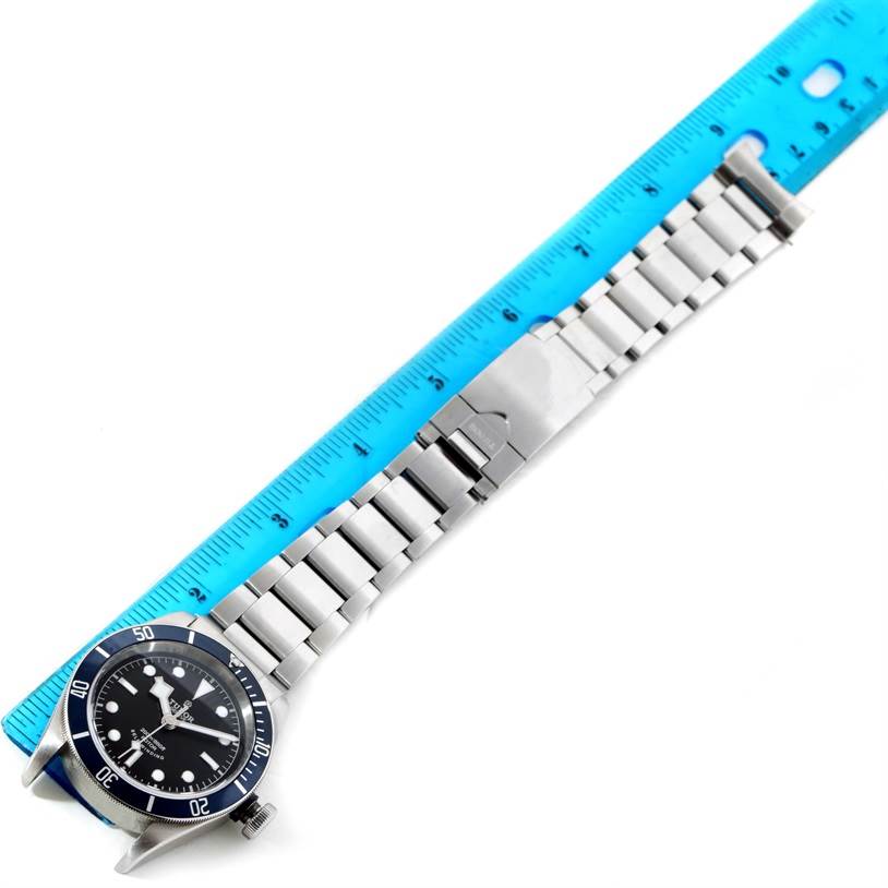 The Tudor Heritage Black Bay 79220 Men's Stainless Steel Black Dial 79220 Men's Stainless Steel Black Dial watch is shown from a top angle, displaying its face, bezel, and metal bracelet beside a ruler.