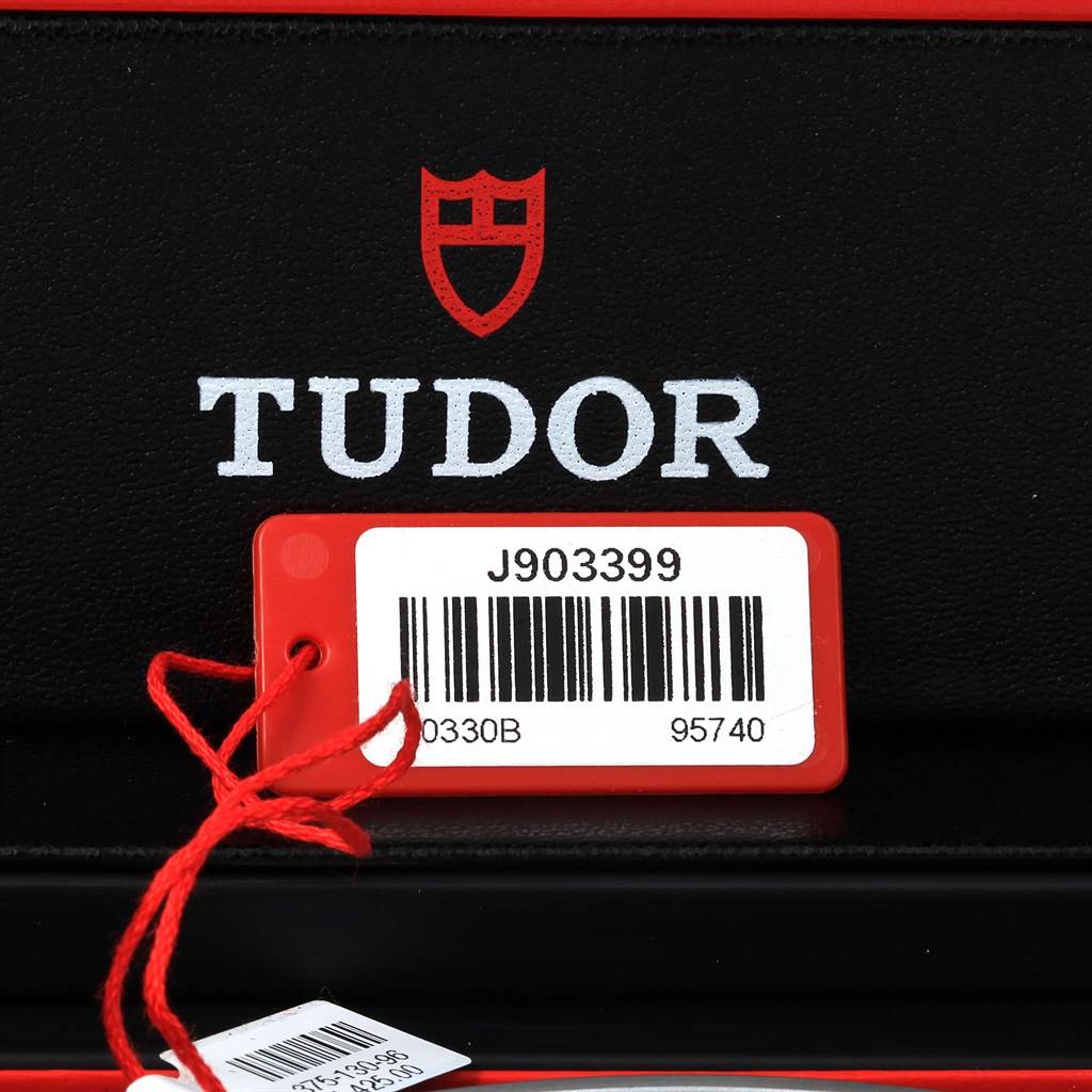 The image shows a tag with the serial number and a close-up of the Tudor logo on packaging, part of the Heritage Chrono watch accessories.