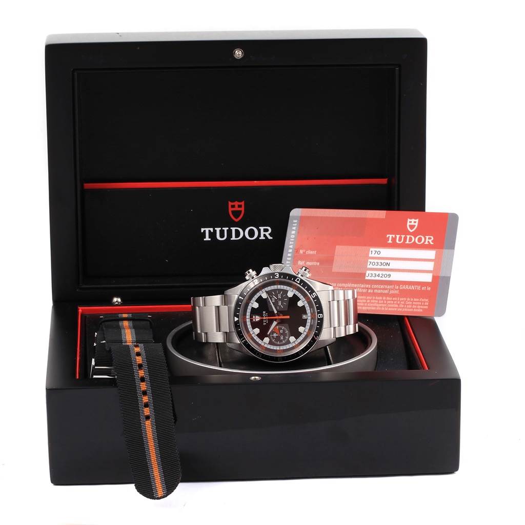 The Tudor Heritage Chrono 70330N Men's Stainless Steel Grey Dial 70330N Men's Stainless Steel Grey Dial watch is shown front-facing in an open box with an additional strap and warranty card displayed.
