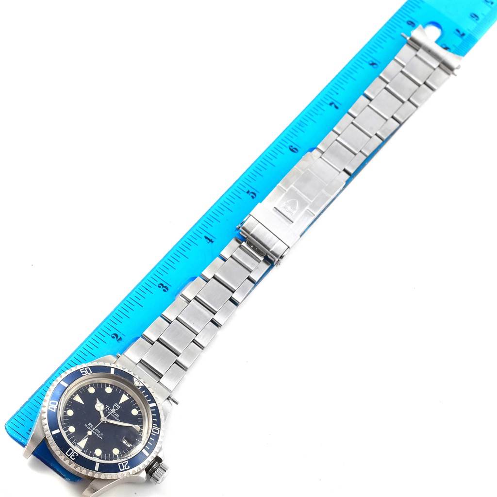 The Tudor Submariner 79090 Men's Stainless Steel Black Dial 79090 Men's Stainless Steel Black Dial watch is shown from a top-down angle, displaying its face, bezel, and metal bracelet beside a ruler.