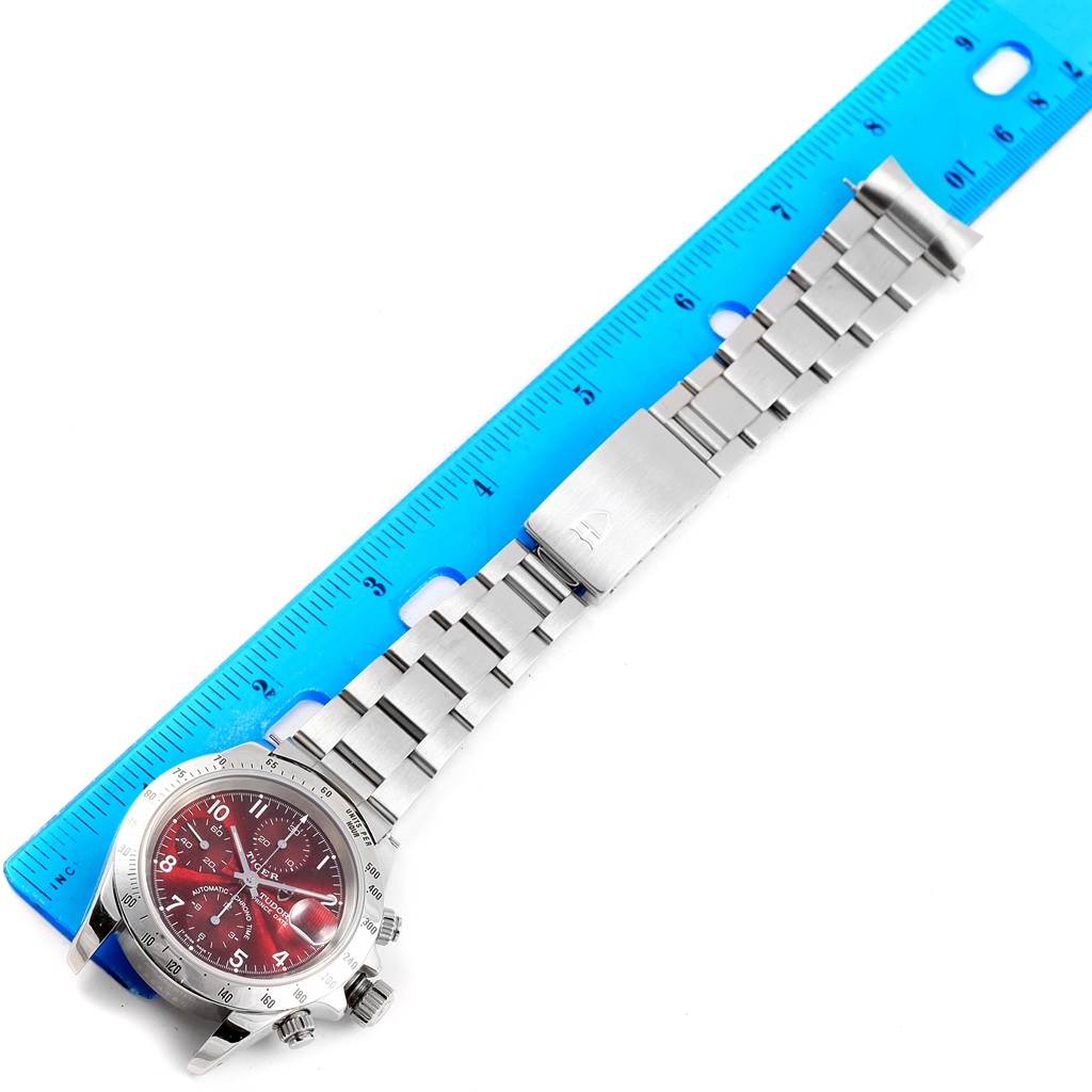 The image shows a Tudor Tiger 79280 Men's Stainless Steel Burgundy Dial 79280 Men's Stainless Steel Burgundy Dial model watch with a red dial and stainless steel bracelet, positioned next to a blue ruler.