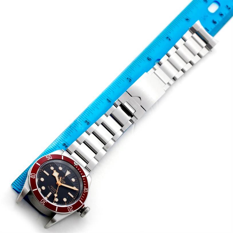 The image shows a top-down view of the Tudor Heritage Black Bay 79220 Men
s Stainless Steel Black Dial 79220 Men
s Stainless Steel Black Dial watch with a metal bracelet next to a blue ruler.