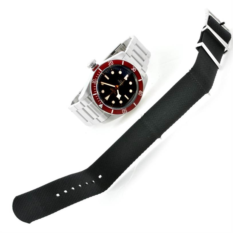 The image shows a Tudor Heritage Black Bay 79220 Men
s Stainless Steel Black Dial 79220 Men
s Stainless Steel Black Dial watch with a partial view of its metal bracelet and a separate black watch strap.