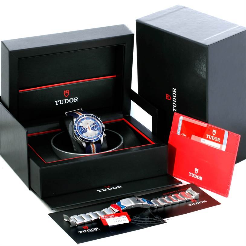 The image shows a Tudor Heritage Chrono 79220R Men's Stainless Steel Blue Dial 79220R Men's Stainless Steel Blue Dial watch at an angle inside its box, accompanied by extra straps, booklets, and packaging.