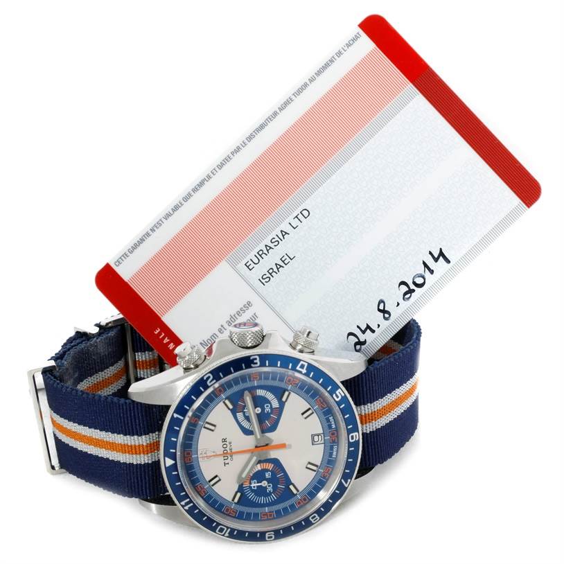 The Tudor Heritage Chrono 79220R Men's Stainless Steel Blue Dial 79220R Men's Stainless Steel Blue Dial watch is shown at an angle, highlighting its face, bezel, and striped fabric strap beside a warranty card.